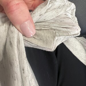 May include: Close-up of a light gray, marled fabric, possibly a t-shirt or other garment. The fabric has a soft, slightly textured appearance with darker flecks throughout. The edge of the fabric is visible, showing a folded seam.