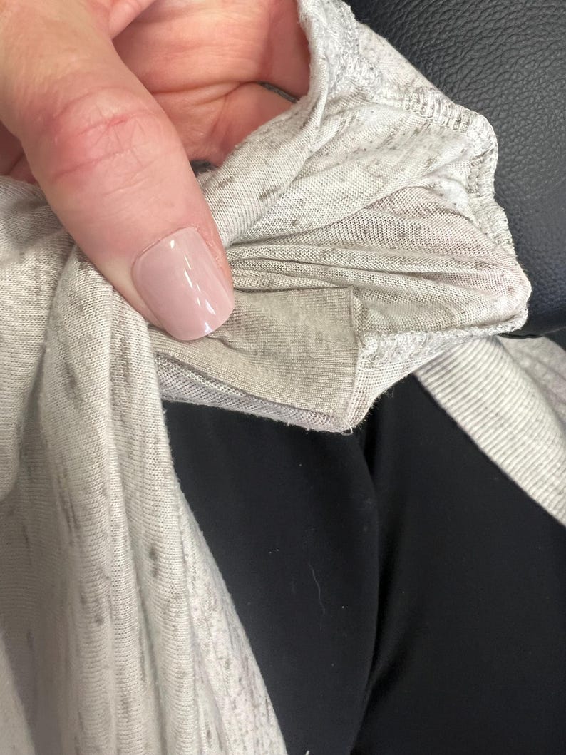 May include: Close-up of a light gray, marled fabric, possibly a t-shirt or other garment. The fabric has a soft, slightly textured appearance with darker flecks throughout. The edge of the fabric is visible, showing a folded seam.