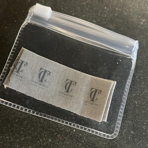May include: A clear plastic zip bag containing a folded gray fabric label. The label has a repeating pattern of the letter "T" with the text "TASCCOMPANY" printed below each letter in a darker gray color.
