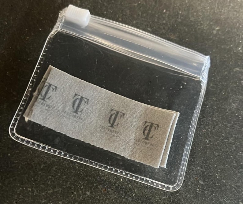 May include: A clear plastic zip bag containing a folded gray fabric label. The label has a repeating pattern of the letter "T" with the text "TASCCOMPANY" printed below each letter in a darker gray color.
