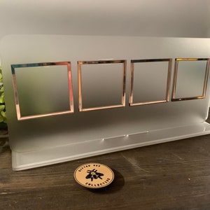Puede incluir: Un expositor de fotos de acrílico transparente con cuatro aberturas cuadradas enmarcadas en oro rosa. El expositor tiene una base para sostenerse y una pequeña pegatina redonda con un logotipo de abeja. El expositor está sobre una superficie de madera.