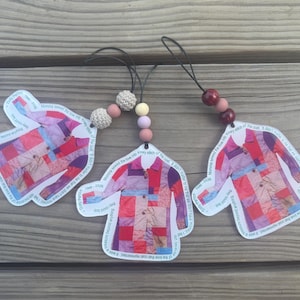 May include: Three coat-shaped ornaments featuring a colorful patchwork pattern in shades of red, pink, and purple. Each ornament includes a black hanging cord with decorative beads and text printed along the edges of the coat design.