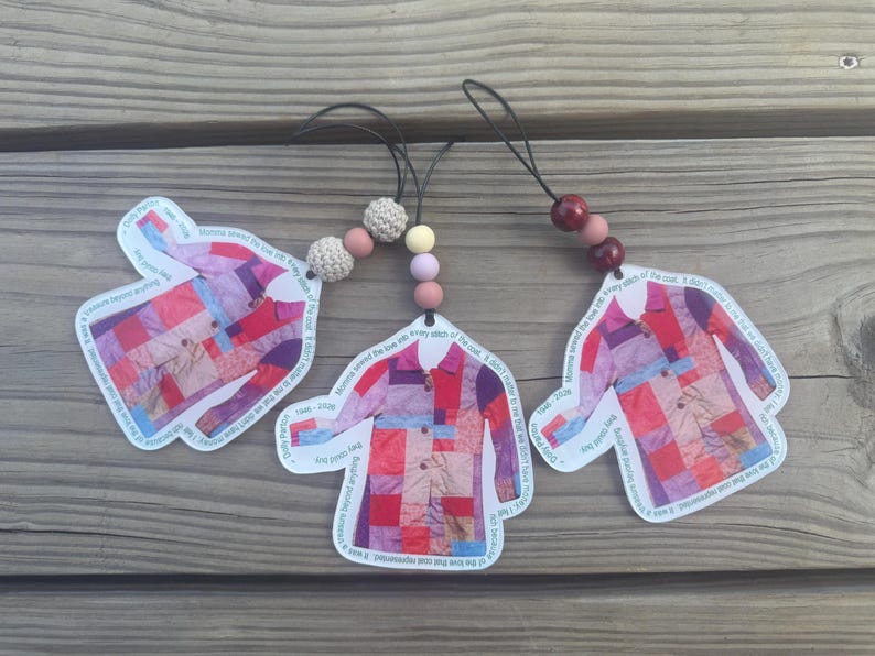 May include: Three coat-shaped ornaments featuring a colorful patchwork pattern in shades of red, pink, and purple. Each ornament includes a black hanging cord with decorative beads and text printed along the edges of the coat design.