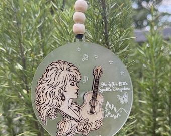 May include: A circular frosted acrylic ornament featuring a laser-engraved profile of a woman with voluminous hair, an acoustic guitar, a butterfly, stars, and musical notes. The text reads Dolly, 1946-2026, and She Left a Little Sparkle Everywhere.