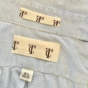 May include: Close-up of a light blue and white striped dress shirt. The shirt has a label with the text "T" repeated multiple times. The size tag reads "16 1/2 34/35".