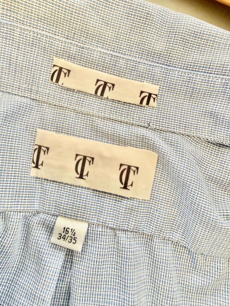 May include: Close-up of a light blue and white striped dress shirt. The shirt has a label with the text "T" repeated multiple times. The size tag reads "16 1/2 34/35".