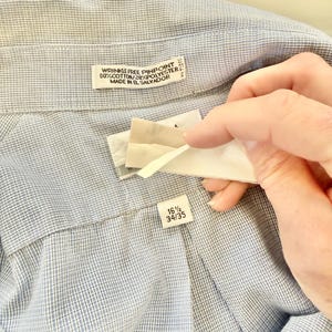 May include: Close-up of a light blue dress shirt with a textured pattern. The shirt has a label that reads "WRINKLE FREE PINPOINT 80% COTTON/20% POLYESTER MADE IN EL SALVADOR." Another label indicates a size of 16 1/2 and 34/35.