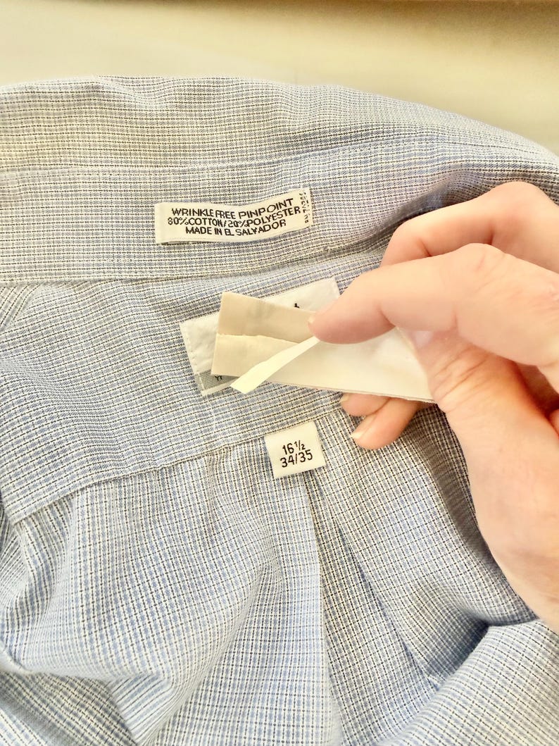 May include: Close-up of a light blue dress shirt with a textured pattern. The shirt has a label that reads "WRINKLE FREE PINPOINT 80% COTTON/20% POLYESTER MADE IN EL SALVADOR." Another label indicates a size of 16 1/2 and 34/35.