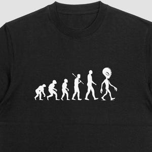 May include: Black crew neck t-shirt featuring a white graphic of the evolution of man, culminating in an alien figure. The design shows a progression from ape-like forms to a humanoid alien. The shirt is made of a soft material.