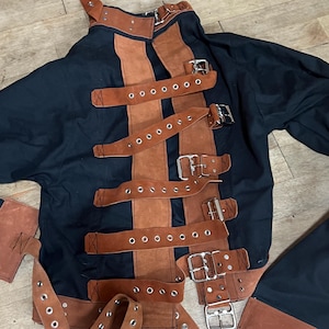 May include: Black and brown straight jacket with multiple leather straps and metal buckles. The jacket has long sleeves and brown leather accents on the shoulders and cuffs. The straps are brown and run vertically across the front.