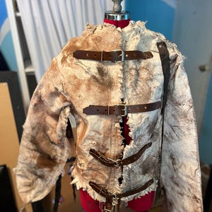 Può includere: Una giacca in pelle bianca sporca e marrone, usurata, con cinturini in pelle marrone e fibbie metalliche. La giacca è esposta su un manichino rosso. La giacca ha un aspetto strappato e usurato.