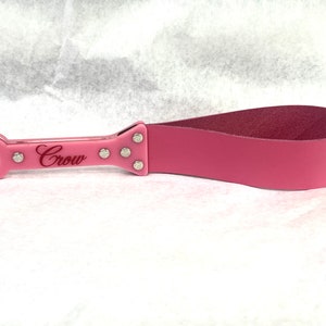 Puede incluir: Paleta de cuero rosa con mango rosa. El mango tiene la palabra "Crow" y adornos plateados. La paleta tiene un lazo en el extremo del mango. El cuero es de color rosa intenso.