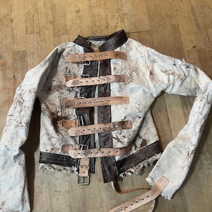 Può includere: Una giacca bianca invecchiata con dettagli marroni e cinturini in pelle. La giacca ha un collo alto, una chiusura a zip e diversi cinturini in pelle con fibbie metalliche. Le maniche sono lunghe e il tessuto sembra usurato.