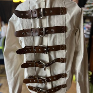 May include: A long-sleeved off-white canvas jacket featuring a vertical row of six brown leather straps with silver-toned metal buckles. The straps are secured over light-colored fabric webbing, extending down the center of the garment's back.