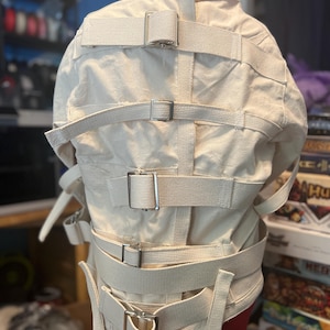 May include: An off-white canvas straightjacket featuring long sleeves and multiple horizontal fabric straps secured with silver-toned metal buckles. The garment is displayed on a mannequin, showing a central vertical strap and various adjustable fasteners.