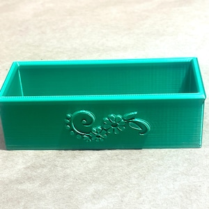 May include: A small, rectangular, green plastic box with a decorative floral design on the front.