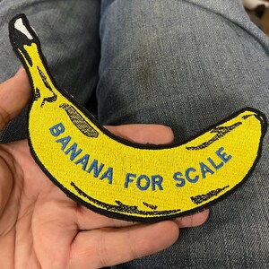 May include: Embroidered yellow banana patch with black outline and the text "BANANA FOR SCALE" in blue. The patch is held against a background of blue denim jeans.