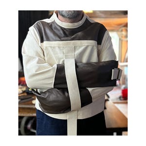 May include: A long-sleeved garment featuring a cream-colored canvas body with dark brown leather panels on the chest and sleeves. The design includes wide, cream-colored fabric straps that cross the front and secure with metal buckles.