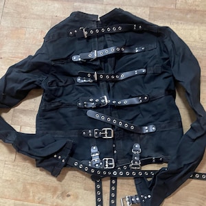May include: Black jacket with multiple leather straps and metal buckles. The jacket has long sleeves and is made of a dark fabric. The straps are secured with metal rivets and buckles, creating a unique and edgy design.
