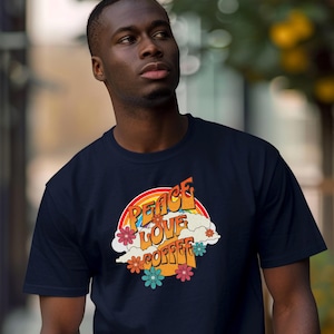 Könnte beinhalten: Marineblaues T-Shirt mit Retro-Design mit den Worten "Peace Love Coffee" in Orange und Gelb, vor einem Regenbogen und Wolken. Das Design beinhaltet bunte Blumen.