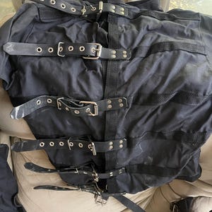 Può includere: Camicia di forza nera in tela con più cinturini in pelle e fibbie in metallo. La giacca ha diversi cinturini orizzontali sul petto e sul busto, con cinturini aggiuntivi per le braccia. La giacca è progettata per limitare i movimenti.