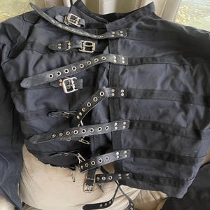 Può includere: Camicia di forza nera con più cinturini in pelle e fibbie metalliche. La giacca ha maniche lunghe ed è realizzata in un tessuto scuro e robusto. I cinturini sono fissati con fibbie metalliche.