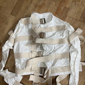 Può includere: Una camicia di forza bianca con multiple cinghie beige e fibbie metalliche. La giacca è realizzata in un materiale liscio, forse simile al raso. Le cinghie sono fissate sul petto, in vita e sulle braccia.