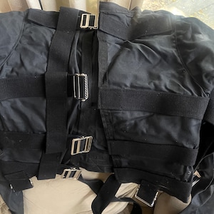 Può includere: Camicia di forza nera con più cinghie nere e fibbie argentate. La giacca è realizzata in un materiale liscio, forse nylon. Le cinghie sono larghe e fissano la giacca. La giacca è appoggiata su una superficie beige.
