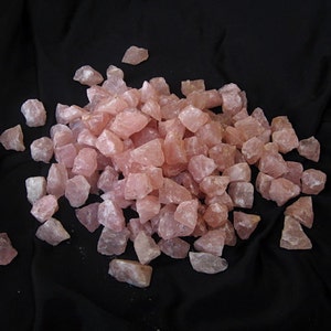 May include: A large pile of pink rose quartz crystals, tumbled and rough, on a black fabric background.