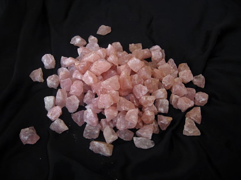 May include: A large pile of pink rose quartz crystals, tumbled and rough, on a black fabric background.