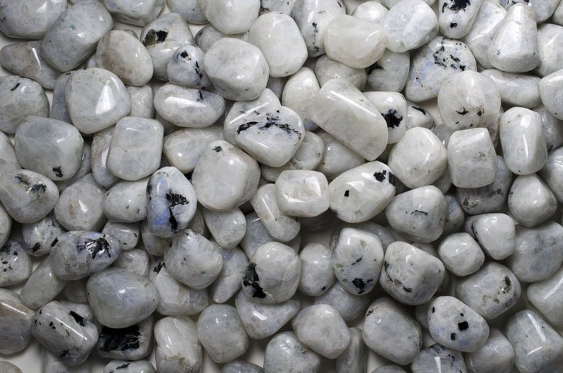 May include: A collection of white moonstone tumbled stones with black inclusions. The stones are smooth and polished, and they have a variety of shapes and sizes.
