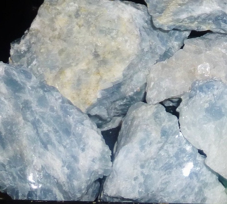 May include: A collection of rough, light blue celestite crystals. The crystals have a translucent, milky appearance and are arranged in a pile.