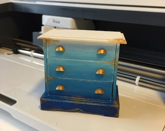 May include: A miniature wooden chest of drawers with three drawers, painted in shades of blue with gold accents. The top is white with gold trim, and each drawer has a gold-colored, half-moon handle. The chest is a decorative item.