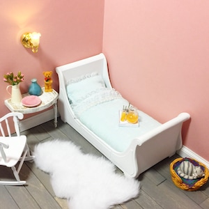 May include: A miniature bedroom scene with a white sleigh bed, a small side table, and a white rocking chair. The bed has a light blue comforter and pillow with white lace trim. A fluffy white rug and a cat bed with a sleeping cat are on the floor.