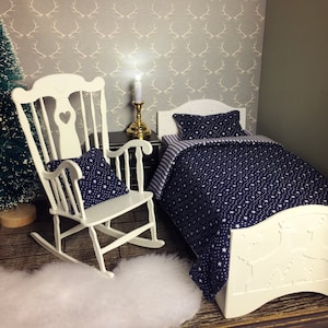 May include: A miniature bedroom scene featuring a white rocking chair with a star-patterned pillow, a bed with a navy blue star-patterned comforter, and a matching pillow. The room has a gray antler wallpaper and a small Christmas tree.