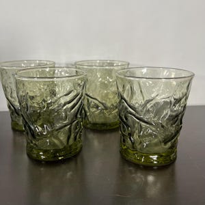 May include: A set of four short drinking glasses made of translucent olive green glass. Each glass features a distinct, irregular, and textured surface pattern that resembles crumpled or melted glass, creating a unique light reflection.