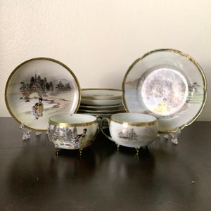 May include: A vintage Japanese porcelain tea set featuring hand-painted scenes of geishas and landscapes. The set includes two teacups with small feet, two plates, and a stack of smaller plates. Gold trim accents the edges of the cups and plates.