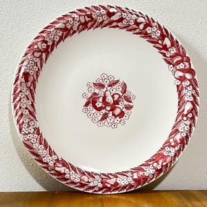 May include: A white ceramic plate with a red fruit and floral design. The rim features a repeating pattern of red fruit and white flowers. The center of the plate has a cluster of red fruit and white flowers.