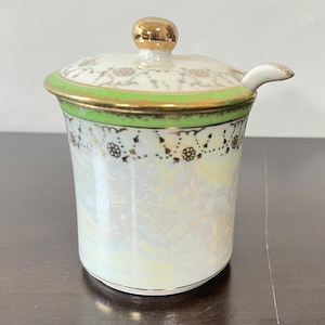 May include: A porcelain condiment jar with an iridescent white finish, featuring a bright green band around the top rim and a gold-trimmed lid with a matching gold knob. A small porcelain spoon rests in the side notch of the lid.