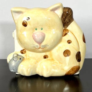 May include: A ceramic cat figurine with a light yellow base color and brown spots. The cat has a pink nose, green eyes, and a gray mouse figurine. The cat is holding a small white object.
