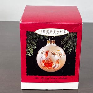 May include: A Keepsake Ornament box with a red exterior. The box features a Christmas ornament with a Peter Rabbit design. The ornament is white with illustrations of Peter Rabbit and other characters. The text "The Tale of Peter Rabbit" is visible.