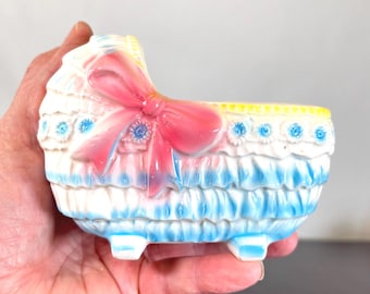 May include: A vintage, pastel-colored baby cradle planter. The cradle features a pink bow, blue and white ruffled trim, and blue floral accents. The planter is made of a glossy material and has small feet.
