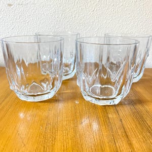May include: Four clear, faceted drinking glasses arranged on a wooden surface. The glasses have a short, cylindrical shape with vertical grooves and a decorative base. The glasses are empty and appear to be made of glass.