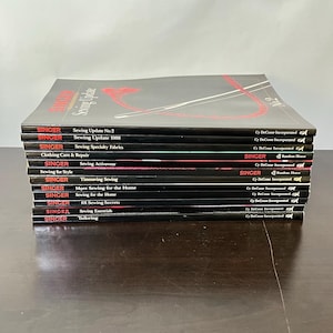May include: A stack of twelve black Singer sewing reference books with white and red text on the spines. Titles include Sewing Update No. 2, Sewing Update 1988, Sewing Specialty Fabrics, Clothing Care & Repair, Sewing Activewear, Sewing for Style, and Tailoring.