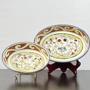 May include: Two oval ceramic dishes featuring a cream-colored center with hand-painted yellow flowers, green foliage, and small red and black berries. The wide rims are decorated with a brown and tan swirling pattern and a dark grey outer edge.