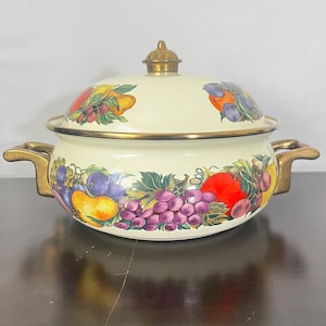 May include: Cream-colored enamelware pot with a lid, adorned with a vibrant fruit design featuring grapes, plums, pears, and tomatoes. The pot has gold-colored handles and trim, and a gold knob on the lid. A decorative kitchenware item.