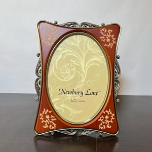 May include: An oval picture frame with a dark wood finish and gold floral accents. The frame features ornate silver-toned metal edges and a central insert printed with the text Newbury Lane and 5x7in 13x18cm.