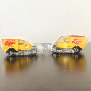 May include: Two yellow toy delivery trucks with red roofs and the Eggo logo. The trucks are in plastic packaging, and have black wheels. The trucks are on a reflective surface.