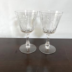 May include: Two clear crystal wine glasses with starburst and vertical line designs. The glasses have a flared bowl, a detailed stem, and a round base. The glasses are on a dark brown surface.