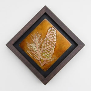 Op de afbeelding: Een ingelijst kunstwerk met een koperen en groene dennenappel met een tak, tegen een warme oranje achtergrond.